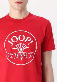 T-shirt en coton rouge avec un grand design graphique blanc portant le texte "JOOP!" et "JEANS", encadré dans un bord circulaire avec des accents floraux.