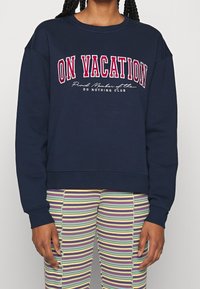 Person som bär en marinblå sweatshirt med texten "PÅ SEMESTER" och randiga byxor i flera färger med vågiga sömmar, stående mot en neutral bakgrund.