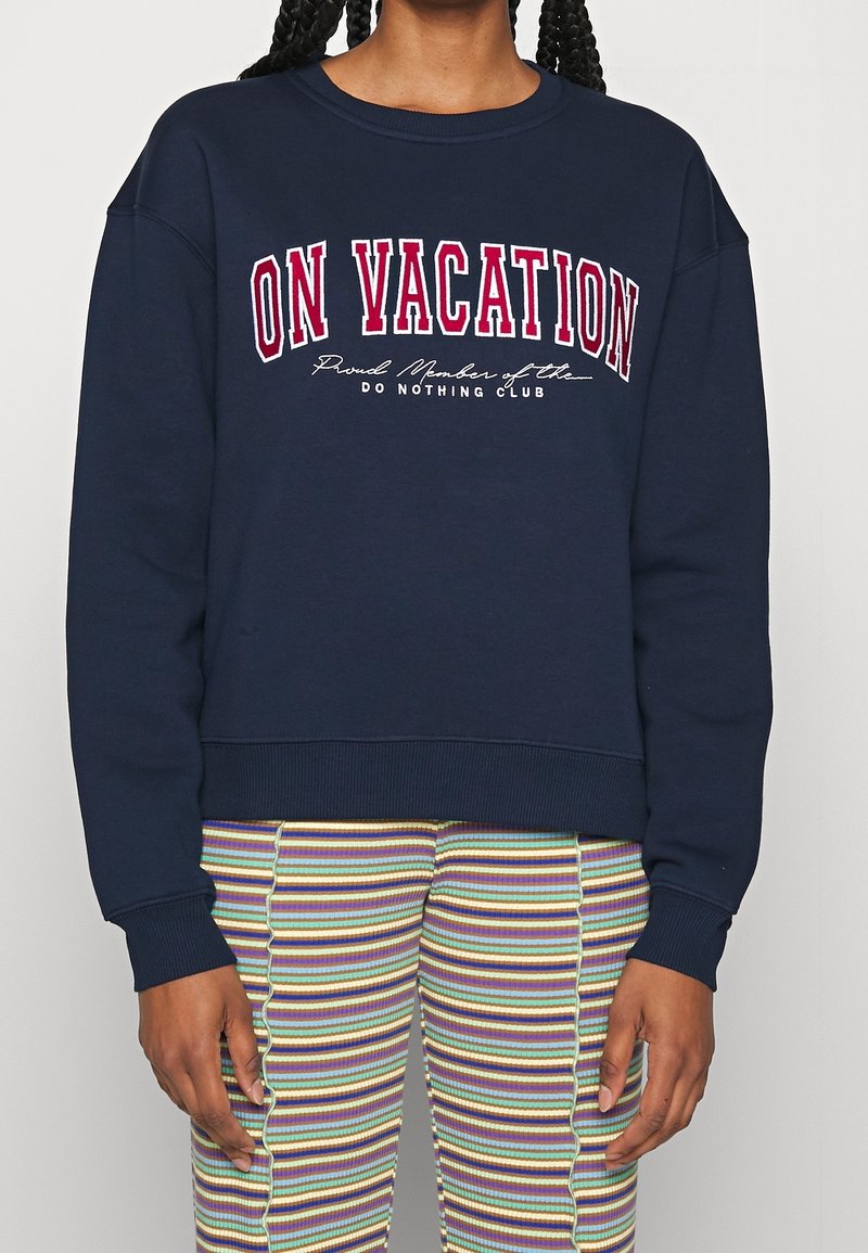 Person som bär en marinblå sweatshirt med texten "PÅ SEMESTER" och randiga byxor i flera färger med vågiga sömmar, stående mot en neutral bakgrund.