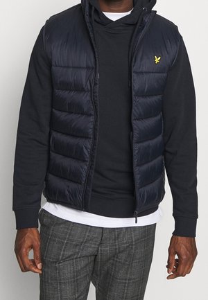 Hombre con chaleco acolchado negro con logo amarillo sobre sudadera negra y camisa blanca, combinado con pantalones gris a cuadros, de pie contra un fondo claro.