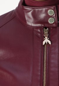 Giacca in pelle bordeaux con colletto rialzato, caratterizzata da due bottoni a pressione e una zip dorata con un design unico del tirante. Texture liscia.