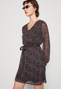 Vestido preto floral com mangas longas e transparentes, decote em V e cintura ajustada. Apresenta pequenas flores rosas, azuis e amarelas num fundo escuro.