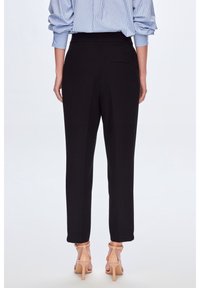 Femme portant un pantalon noir ajusté, une chemise à rayures bleu clair à manches longues et des sandales à talons nude, vue de dos sur fond blanc.