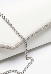 Λευκή ψεύτικη δερμάτινη τσάντα clutch με ασημένια αλυσίδα, διαθέτοντας υφή στην άκρη που κοσμείται από μικρούς κρυστάλλους κατά μήκος της γραμμής κλεισίματος.