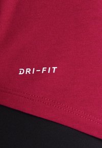 Tessuto rosso con logo "DRI-FIT" bianco. Texture liscia e design a maniche corte visibili, con cucitura nera a contrasto in basso.