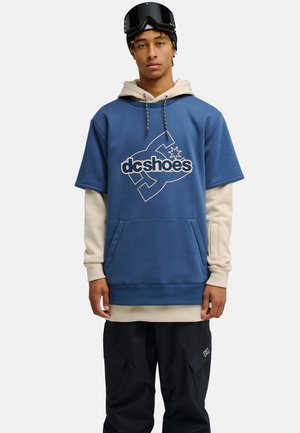 Młody mężczyzna ubrany w niebieską, z krótkim rękawem bluzę DC Shoes na beżową bluzkę z długim rękawem, czarne spodnie oraz czarne gogle narciarskie na głowie.