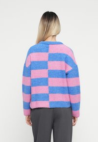 Ženska z dolgimi blond lasmi, oblečena v potemnjen pulover z debelo pletenico, ki ima izmenične modre in roza horizontalne črte, prikazana s hrbta.