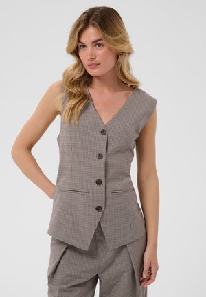 Frau mit langen, welligen, blonden Haaren trägt eine maßgeschneiderte, ärmellose, karierte Weste mit braunen Knöpfen und eine dazu passende hoch taillierte Hose, steht selbstbewusst.