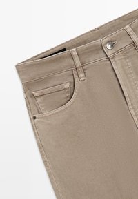 Gros plan sur un pantalon beige montrant la ceinture, le bouton, les passants et les poches avant sur fond blanc.