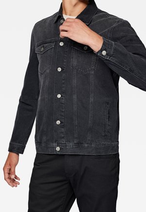 Homme ajustant le col d'une veste en jean noire avec des boutons argentés, associée à un pantalon noir, sur fond blanc.