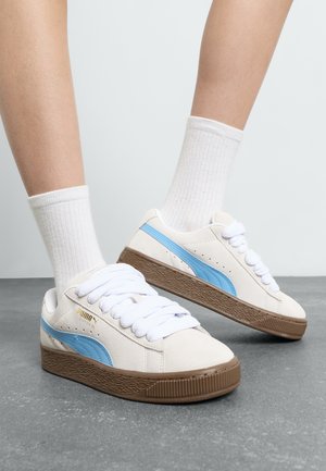 Le sneakers Puma in suede presentano una tomaia bianca, dettagli azzurri chiari e una suola in gomma marrone texturizzata, con lacci bianchi spessi.