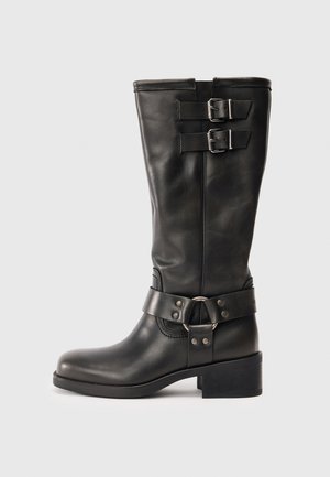 Bota de cuero negra hasta la mitad de la pantorrilla con dos hebillas plateadas cerca de la parte superior y una correa decorativa con anillo de metal alrededor del tobillo, con tacón cuadrado.