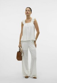 Witte top met ruches en een gathered halslijn, gecombineerd met wijduitlopende, gestreepte witte broeken. Een bruine handtas en bijpassende platte schoenen maken de look af.