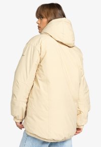 Giacca puffa di colore beige chiaro, dotata di cappuccio, polsini elastici e orlo cinchato. Presenta un logo sulla manica sinistra e una texture liscia.