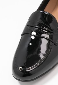 Mocassin en cuir verni noir avec un bout arrondi, une surface lisse et des coutures décoratives sur la partie supérieure. La semelle est plate et légèrement texturée.