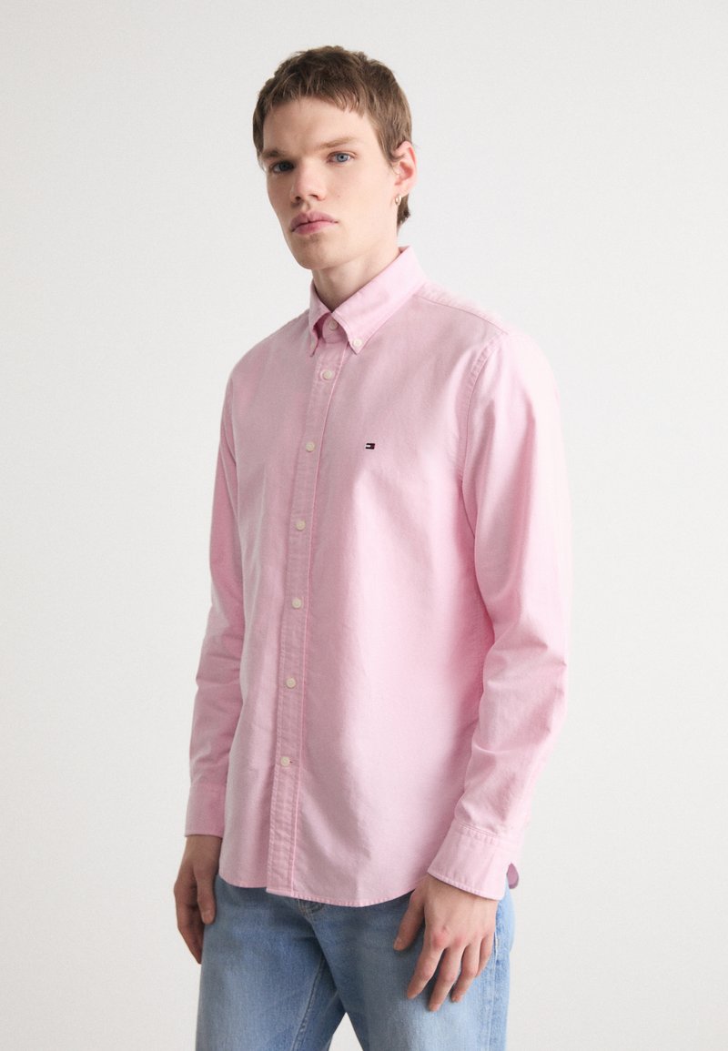 Gaiši rozā garām piedurknēm pogājama krekla ar apkakli, ar mazu logo uz krūtīm un gludu kokvilnas tekstūru.