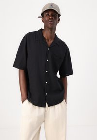 Camisa de hombre negra de manga corta con bolsillo, hecha de una tela texturizada, con un cuello clásico y botones blancos. Se lleva con pantalones beige.