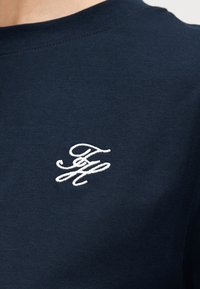 Μπλούζα σε σκούρο μπλε χρώμα, με λαιμόκοψη, με κεντημένα με λευκά γράμματα με καλλιγραφικό στυλ τα αρχικά "TH" στην αριστερή πλευρά του στήθους.