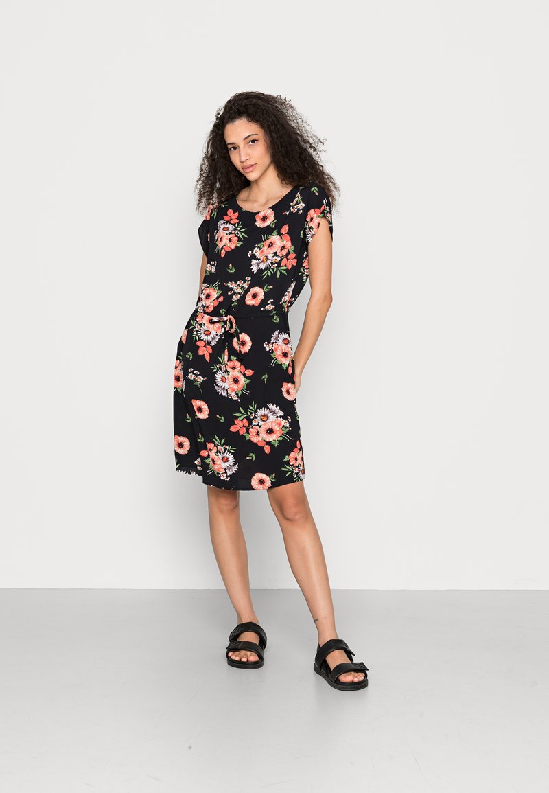 Vestido curto floral em preto com flores laranja e brancas. Mangas curtas, cintura ajustável, até aos joelhos. Usado com sandálias pretas.