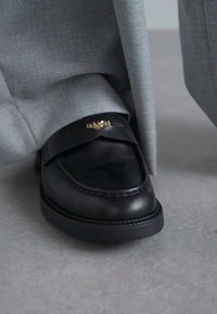 Black leather loafer shoe with a branded gold accent on the strap, paired with gray fabric trousers, resting on a gray surface.