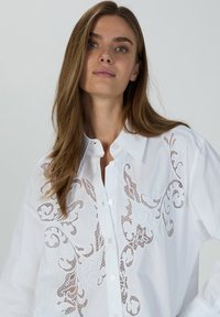 White button-up shirt made of cotton, featuring intricate lace embroidery in a swirling pattern across the front. Smooth texture, relaxed fit.
