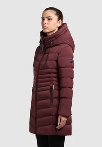 Cappotto imbottito bordeaux con collo alto, cappuccio con coulisse e tasche con cerniera. Presenta un design trapuntato e una silhouette leggermente affusolata.