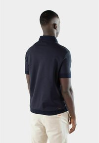 Marineblå poloshirt med korte ermer, teksturert stoff og ribbet krage. Har en liten rød logoakcent nær sømmen.