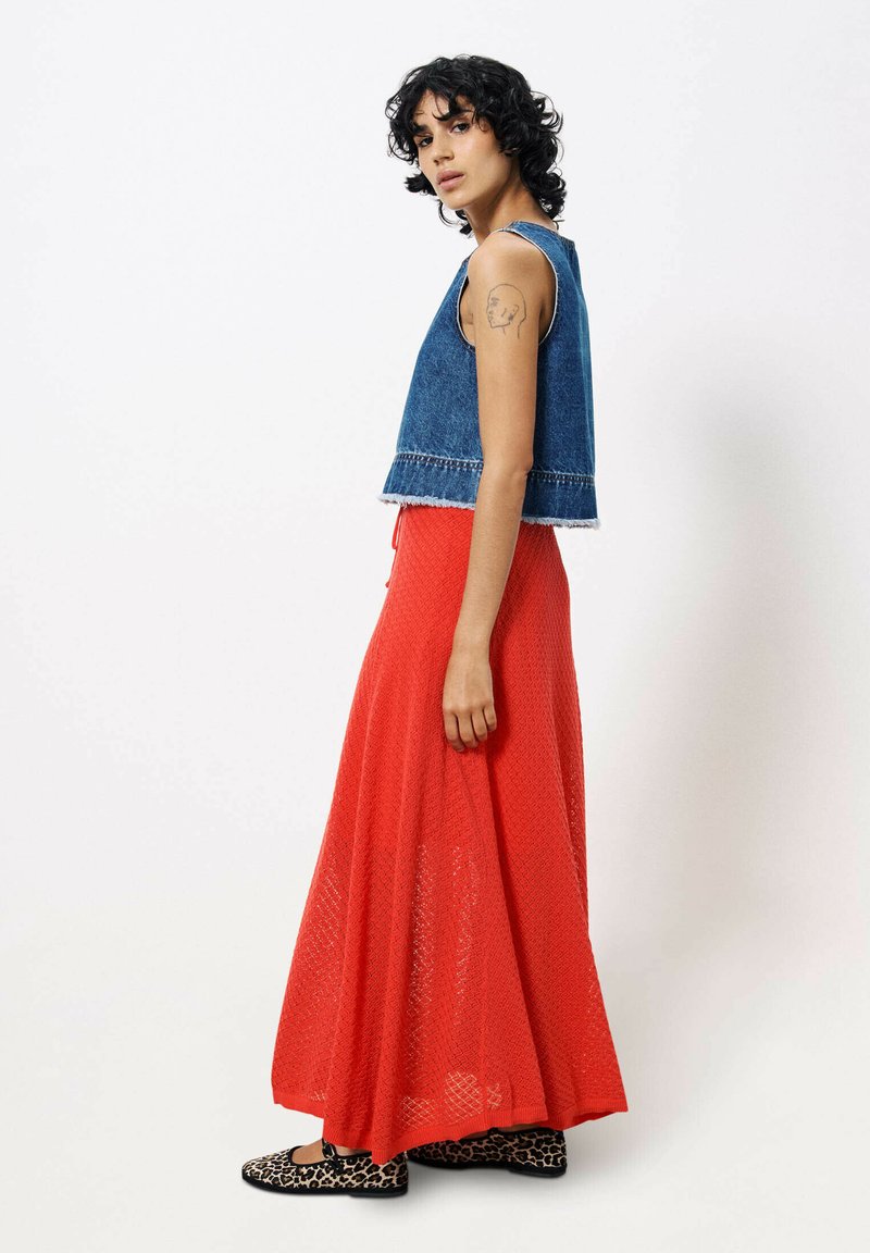 Personne avec des cheveux bouclés portant un haut en denim sans manches, une longue jupe maxi rouge texturée, des ballerines à imprimé léopard et un tatouage sur le bras.