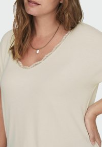Gai beige krāsu V veida dekoltē topiņš ar mežģīņu apdari, mīksts audums, brīvs piegriezums un īsas piedurknes, valkāts ar kārtām ķēdi un pērlēm.