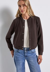 Brun kort jacka i slät tyg, med tryckknappar, ribbade ärmslut och rund halsringning. Bärs över en ljus topp.