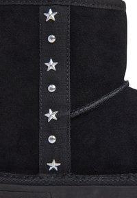 Dettaglio di stivali in suede neri con una cinghia verticale nera decorata da stelle argento alternando e borchie rotonde.