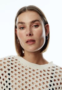 Strickpullover in Creme mit durchbrochenem Muster und schwarzen Akzenten. Verfügt über einen gerippten Halsausschnitt und kurze Ärmel, getragen über einer glatten Textur.