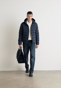 Mornarica puffer jakna s kapuljačom, bijela polo majica, tamne traperice i crna torba. Glatka tekstura s laganim sjajem.