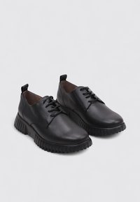 Zapatos de cuero negros con cordones, suelas gruesas y estriadas, diseño de tobillo bajo y tiradores en la parte trasera sobre un fondo gris claro.