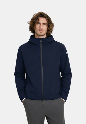 Uomo con capelli ricci che indossa una giacca blu navy con cerniera e cappuccio e pantaloni grigio scuro, con le mani in tasca, rivolto verso avanti.