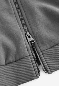 Nahaufnahme eines grauen Stoffes mit einem teilweise geöffneten Metallreißverschluss mit der Aufschrift "ALLSAINTS" und rippenförmigen Saumdetails.