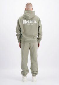 Olijfgroene hoodie en joggingbroek set. Hoodie heeft een groot wit logo op de achterkant, geribbelde manchetten en een kangoeroezak. Gladde textuur, comfortabele pasvorm.