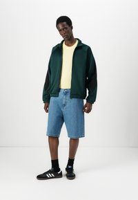 Πράσινο και μαύρο ζακέτα με φερμουάρ, κίτρινη μπλούζα, μπλε τζην σορτς και μαύρα αθλητικά παπούτσια με λευκές ρίγες; εμφανισμένο casual ντύσιμο.