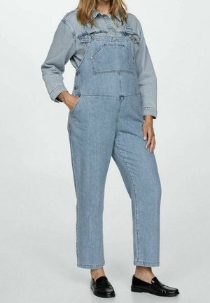 Kvinde iført lyseblå denimoveralls med en matchende denimskjorte og sorte loafers, stående med den ene hånd i lommen.