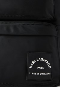 Musta nailoniselkäre, jossa on suorakulmainen muoto, sileä pinta ja näkyvä valkoinen logo. Mukana tyylikkäät vetskarit.