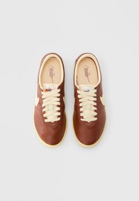 Brązowe skórzane trampki z kremowymi sznurowadłami, zaokrąglonym noskiem i kontrastowym białym logo w kształcie znaku. Miękka wewnętrzna wyściółka i teksturowana gumowa podeszwa.