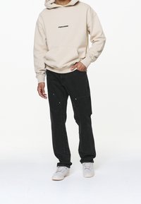 Sudadera con capucha beige con el logo "PEGADOR", jeans cargo negros con bolsillos laterales, zapatillas grises, corte holgado, tela lisa y diseño minimalista.