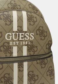 Zaino marrone con il logo "GUESS" e il testo "EST 1981", decorato con strisce bianche e un motivo intricato. Chiusura con cerniera visibile.