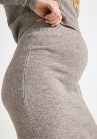 En sidovy av en gravid person som försiktigt håller sin tröja, vilket visar deras rundade mage, klädd i en mjuk, beige stickad outfit.