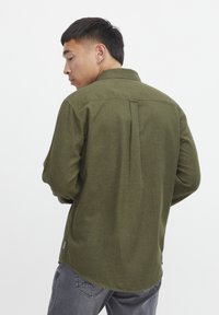 Bărbat cu păr scurt și închis la culoare, îmbrăcat într-o bluză de lungă cu mânecă, de culoare verde măslin, și blugi gri, întors cu spatele, pe un fundal alb simplu.