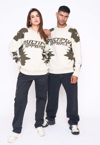 Crème trui met olijfgroen abstract ontwerp en de tekst "MULTIPLY APPAREL", gecombineerd met zwarte cargo-broeken en sneakers.