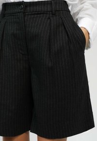 Shorts noirs à rayures avec des plis, fabriqués en tissu texturé. Dotés d'une fermeture à bouton et de poches latérales. Coupe ajustée avec une longueur mi-cuisse.