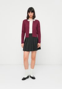 Femme portant un cardigan bordeaux sur une blouse blanche et une jupe plissée noire, se tenant devant un fond blanc uni.
