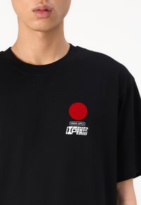 Melnas kokvilnas T-krekls ar sarkanu apli un baltu teksta logo kreisajā krūšu daļā. Īsās piedurknes, apaļa kakla dizains.
