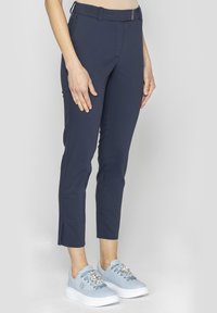 Granatowe, elastyczne, dopasowane spodnie z metalowym zapięciem i rozcięciami na kostkach, w połączeniu z jasnoniebieskimi trampkami z dekoracyjnymi zdobieniami.